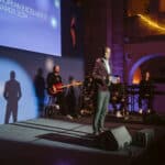 Mann in Anzug spricht auf einer Bühne, umgeben von Musikern, bei den European Excellence Awards 2024.