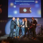 Fünf Männer in Anzügen stehen auf einer Bühne, während eine Auszeichnung überreicht wird. Im Hintergrund ein Bildsch...