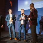 Fünf Männer auf einer Bühne, einer hält einen Preis, während ein anderer spricht; im Hintergrund eine Präsentation.