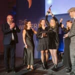 Gruppe von sechs Personen auf einer Bühne, eine Frau hält einen Preis hoch, alle applaudieren und lächeln.