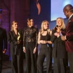 Sechs Personen auf einer Bühne, eine hält ein Mikrofon, andere schauen zu; festliche Atmosphäre mit Beleuchtung.