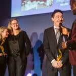 Zwei Preisträger halten Auszeichnungen, während ein Moderator ihnen Fragen stellt. Hintergrund mit Präsentation.