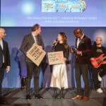 Vier Personen auf einer Bühne, zwei halten Schilder mit politischen Botschaften, im Hintergrund eine Band.