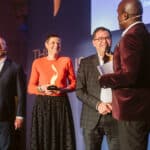 Vier Personen auf einer Bühne bei einer Preisverleihung, eine hält eine Trophäe, alle lächeln und sprechen.