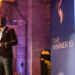 Mann in eleganter Kleidung steht auf einer Bühne und spricht ins Mikrofon, im Hintergrund steht "THE WINNER IS".