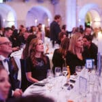 Elegante Veranstaltung mit Gästen an festlich gedeckten Tischen, Kerzenlicht und aufmerksamem Publikum.