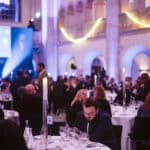 Elegantes Bankett mit gedeckten Tischen, Kerzenlicht und Gästen, die sich unterhalten oder auf ihre Handys schauen.