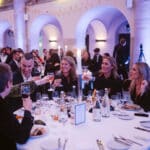 Gruppierung von Gästen bei einer Feier, die mit Gläsern anstoßen, umgeben von festlicher Dekoration und Tischen.