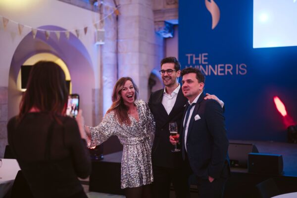 Drei Personen posieren lächelnd vor einer blauen Wand mit dem Text "THE WINNER IS", während eine vierte Person fotografi...