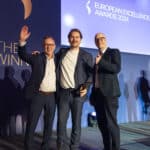 Drei Männer in Anzügen posieren lächelnd auf einer Bühne bei den European Excellence Awards 2024.