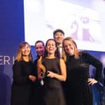 Fünf Personen posieren lächelnd auf einer Bühne, eine hält einen Preis in der Hand, im Hintergrund ein Bildschirm.