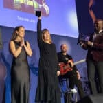Frau in schwarzem Kleid hebt einen Preis auf der Bühne, umgeben von weiteren Personen und einem Bildschirm im Hintergrund.