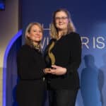 Zwei Frauen in eleganter Kleidung halten gemeinsam einen Preis vor einem blauen Hintergrund mit Schriftzug.