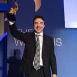 Mann in Anzug hält einen Preis hoch, steht vor einer blauen Wand mit Schriftzug und Schatten.