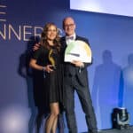 Zwei Personen stehen auf einer Bühne, eine hält einen Preis, der Hintergrund zeigt eine Auszeichnungstafel.