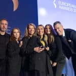 Sechs Personen posieren lächelnd auf einer Bühne, eine hält einen Preis, im Hintergrund das Logo der European Excellenc...
