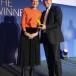 Zwei Personen stehen auf einer Bühne, halten eine Auszeichnung und lächeln, mit einem blauen Hintergrund und Spotlights.