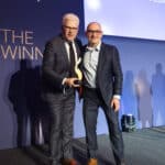 Zwei Männer in Anzügen halten einen Preis auf einer Bühne vor einem Banner mit der Aufschrift "European Excellence Awar...