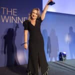 Frau in schwarzem Kleid hält Trophäe hoch, steht auf einer Bühne mit blauer Hintergrundbeleuchtung und Schatten.