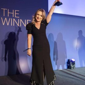 Frau in schwarzem Kleid hält Trophäe hoch, steht auf einer Bühne mit blauer Hintergrundbeleuchtung und Schatten.