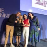 Vier Personen posieren lächelnd auf einer Bühne, eine hält einen Preis in der Hand, im Hintergrund ein Bildschirm.