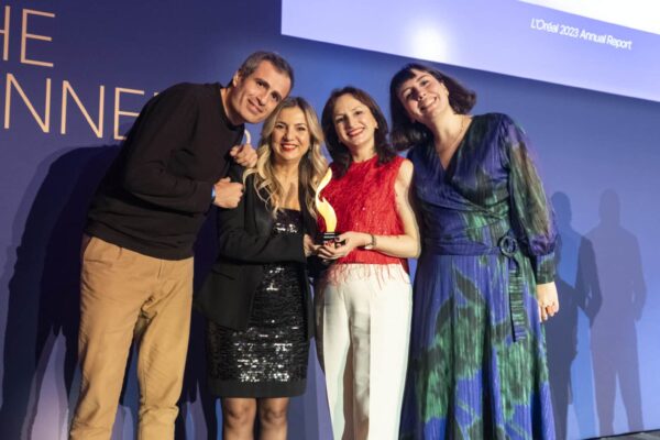 Vier Personen posieren lächelnd auf einer Bühne, eine hält einen Preis in der Hand, im Hintergrund ein Bildschirm.