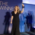 Frau in schwarzem Kleid hält einen Preis hoch, steht vor einer blauen Wand mit dem Text "THE WINNERS". Schatten im Hinter...