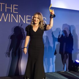 Frau in schwarzem Kleid hält einen Preis hoch, steht vor einer blauen Wand mit dem Text "THE WINNERS". Schatten im Hinter...