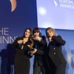 Drei Frauen in eleganter Kleidung posieren mit einer Auszeichnung vor einem blauen Hintergrund mit dem Logo der European A...