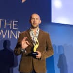 Mann mit braunem Sakko hält einen goldenen Preis in der Hand, steht vor einer blauen Wand mit dem Schriftzug "THE WINNER".