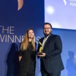 Zwei Personen stehen auf einer Bühne und halten einen Preis, im Hintergrund ein Banner mit dem Text "The Winners".