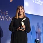 Frau in schwarzem Kleid hält einen Preis, steht vor einer blauen Wand mit Schriftzug "THE WINNER" und "EUROPEAN AWARDS 20...