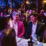 Zwei Männer in eleganter Kleidung sitzen an einem Tisch bei einer festlichen Veranstaltung, umgeben von Gästen und Kerze...