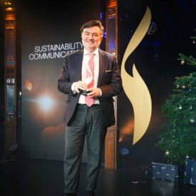 Mann in Anzug steht auf einer Bühne mit Weihnachtsbaum und Dekoration, hält ein Mikrofon, Hintergrund mit Lichtern.