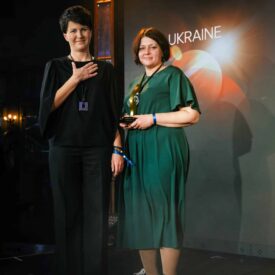 Zwei Frauen stehen auf einer Bühne, eine hält einen Preis, die andere legt die Hand aufs Herz. Hintergrund mit "UKRAINE".