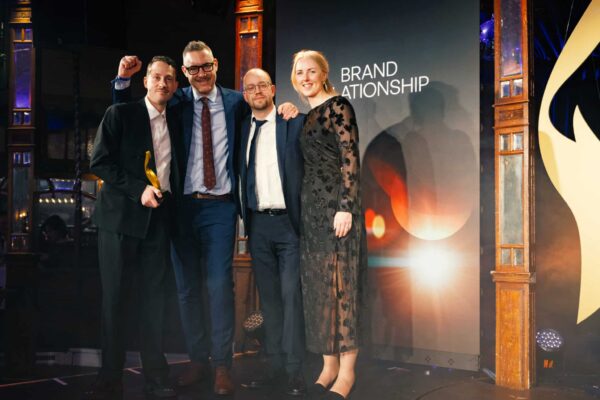 Vier Personen posieren auf einer Bühne, eine hält einen Preis, im Hintergrund ein Banner mit dem Text „Brand Relations...